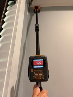 A hand holds a moisture meter on a telescoping pole against intact drywall near the ceiling. The meter display reads 490 in red.